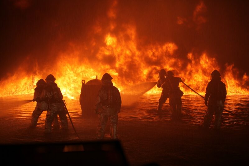 Firefighters in front of a wall of flames, wading through shallow water